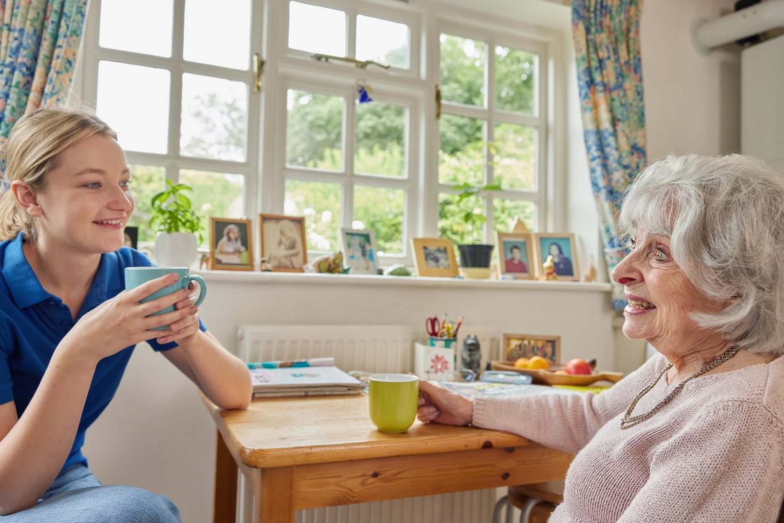 An older woman and her home support worker share a cup of tea and a laugh at her kitchen table.